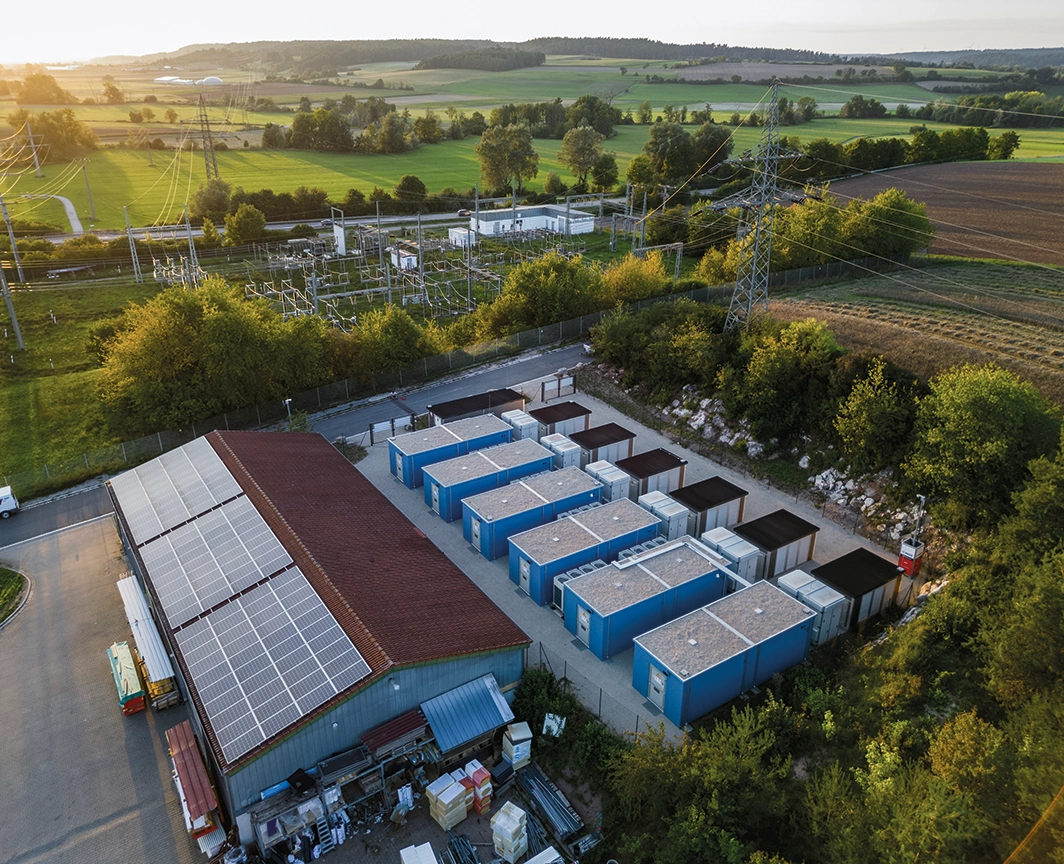 Wir sehen eine Batteriespeicheranlage in mitten von grünen Feldern. Darüber spannen sich Stromnetze. Auf dem Dach einer Lagerhalle vor den Batteriespeichern sind PV Anlagen montiert. Die Abendsonne strahlt im Hintergrund.
